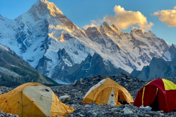 Tents at a high-altitude campsite with a breathtaking view of the Karakoram mountains, including a snow-covered peak glowing under the golden sunset.